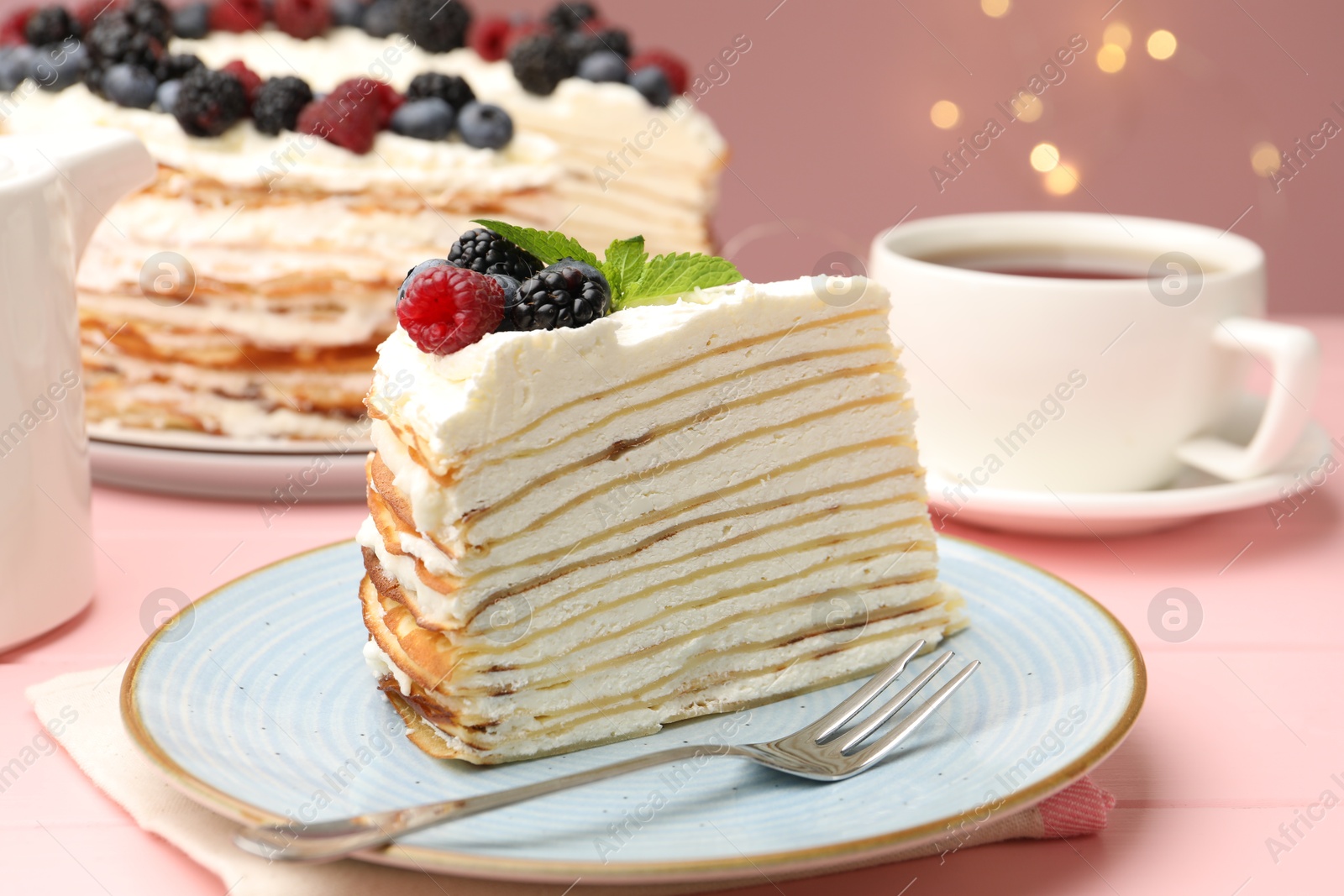 Photo of Piece of tasty crepe cake with berries served on pink wooden table against blurred lights, closeup