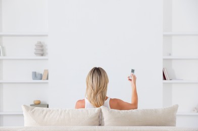 Photo of Woman using remote to control air conditioner on sofa at home, back view