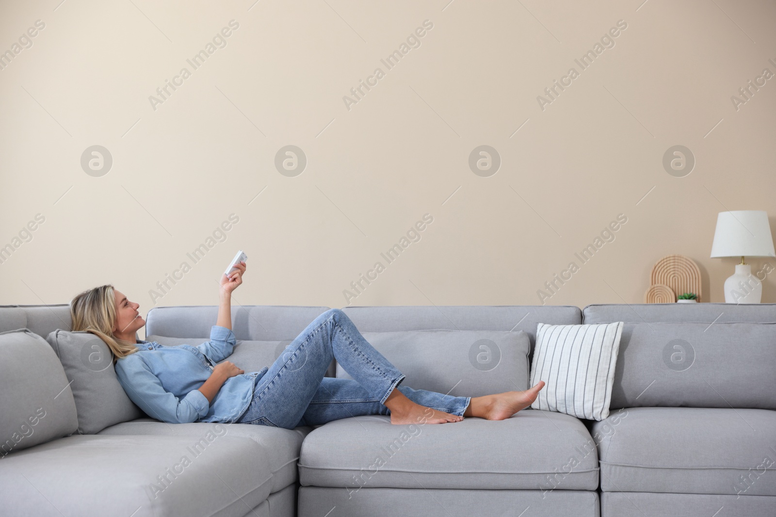 Photo of Woman using remote to control air conditioner on sofa at home