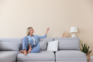 Photo of Woman using remote to control air conditioner on sofa at home