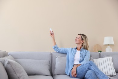 Photo of Woman using remote to control air conditioner on sofa at home