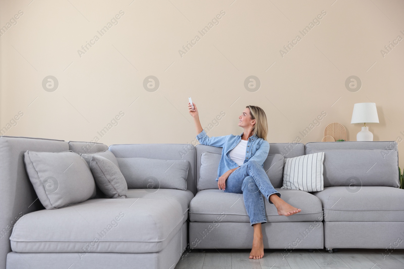Photo of Woman using remote to control air conditioner on sofa at home