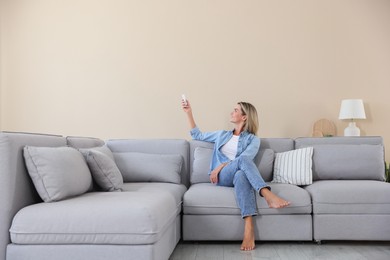 Photo of Woman using remote to control air conditioner on sofa at home
