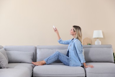 Photo of Woman using remote to control air conditioner on sofa at home