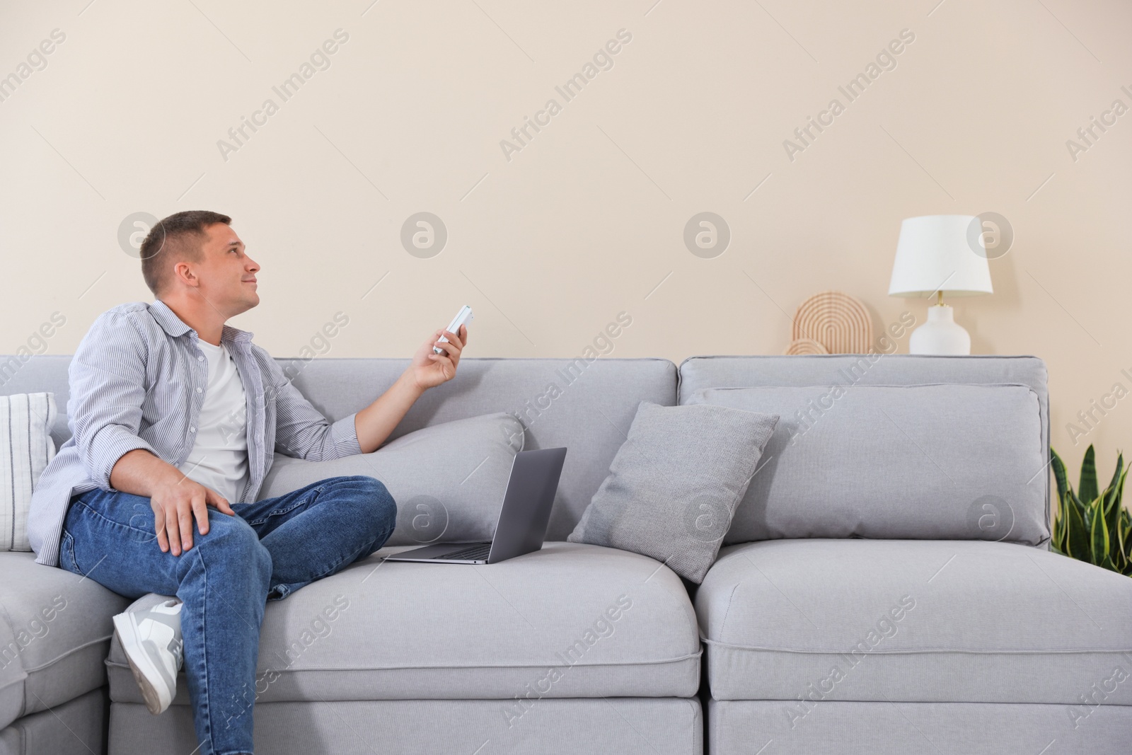 Photo of Man using remote to control air conditioner on sofa at home
