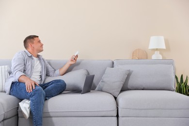 Photo of Man using remote to control air conditioner on sofa at home