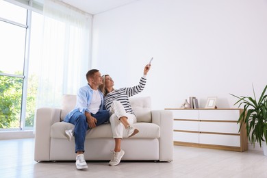 Photo of Man and woman using remote to control air conditioner on sofa at home