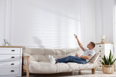 Photo of Man using remote to control air conditioner on sofa at home