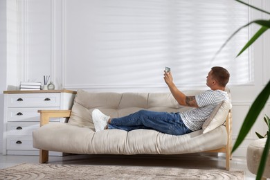 Photo of Man using remote to control air conditioner on sofa at home