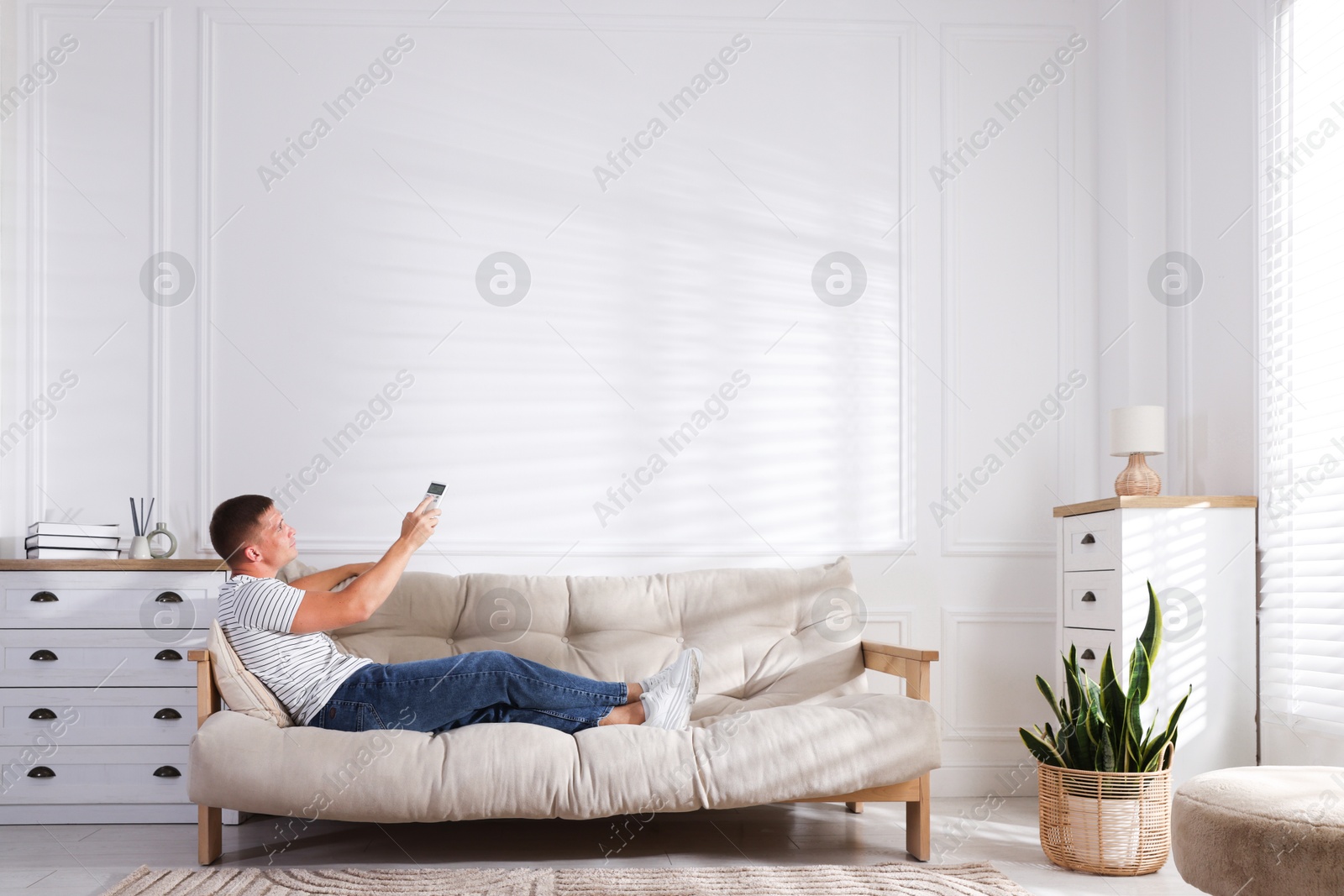 Photo of Man using remote to control air conditioner on sofa at home