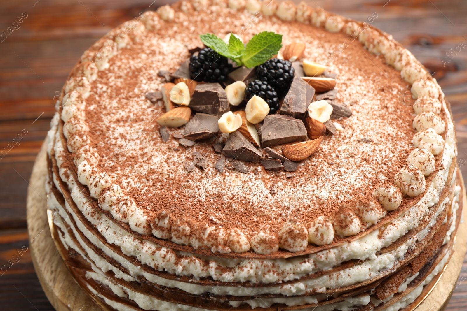 Photo of Delicious crepe cake with chocolate, nuts, blackberries and cocoa powder on wooden table, closeup