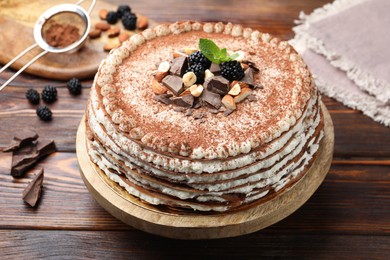 Photo of Delicious crepe cake with chocolate, nuts, blackberries and cocoa powder on wooden table, closeup