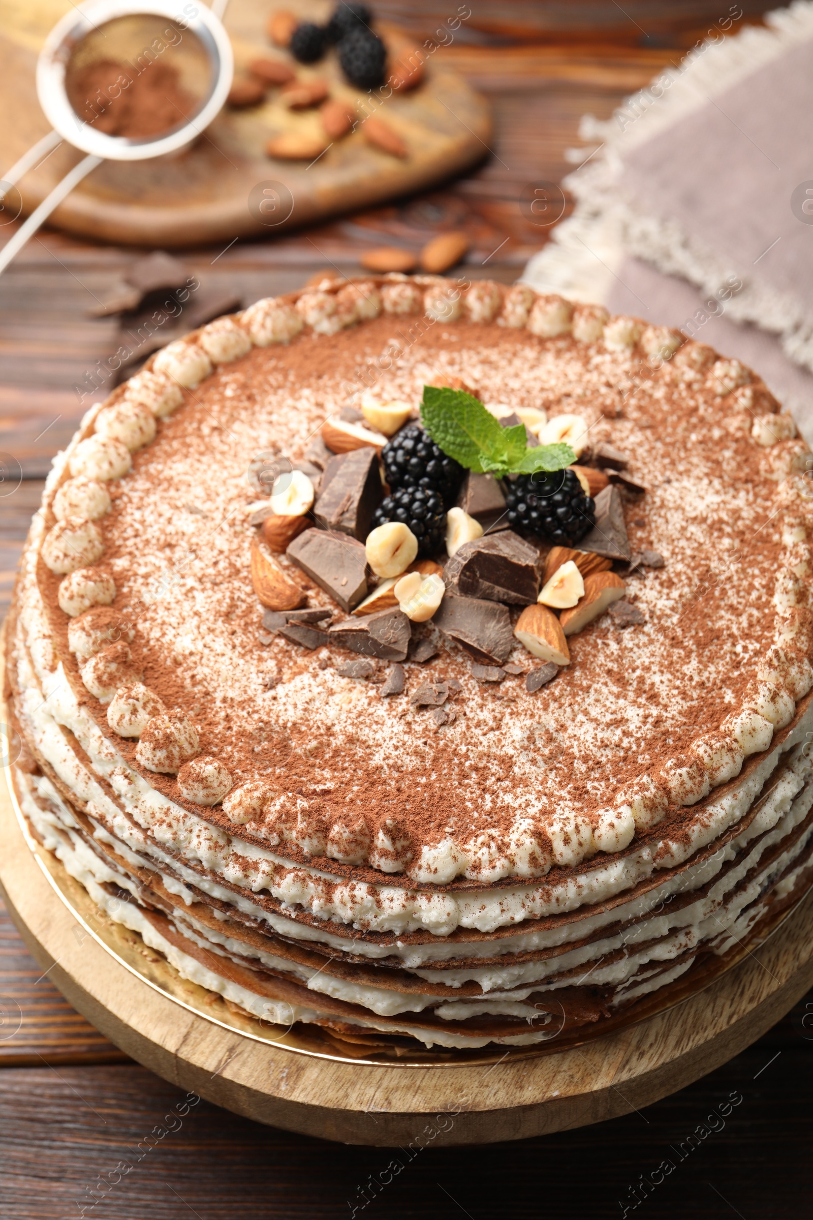 Photo of Delicious crepe cake with chocolate, nuts, blackberries and cocoa powder on wooden table, closeup