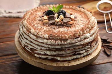 Photo of Delicious crepe cake with chocolate, nuts, blackberries and cocoa powder on wooden table, closeup