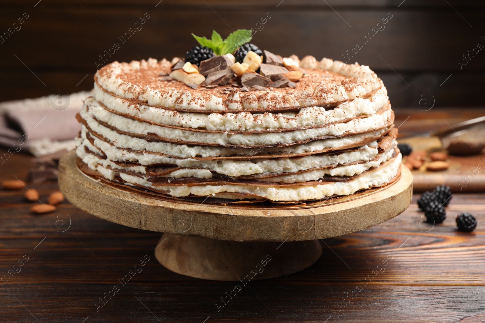 Photo of Delicious crepe cake with chocolate, nuts, blackberries and cocoa powder on wooden table, closeup