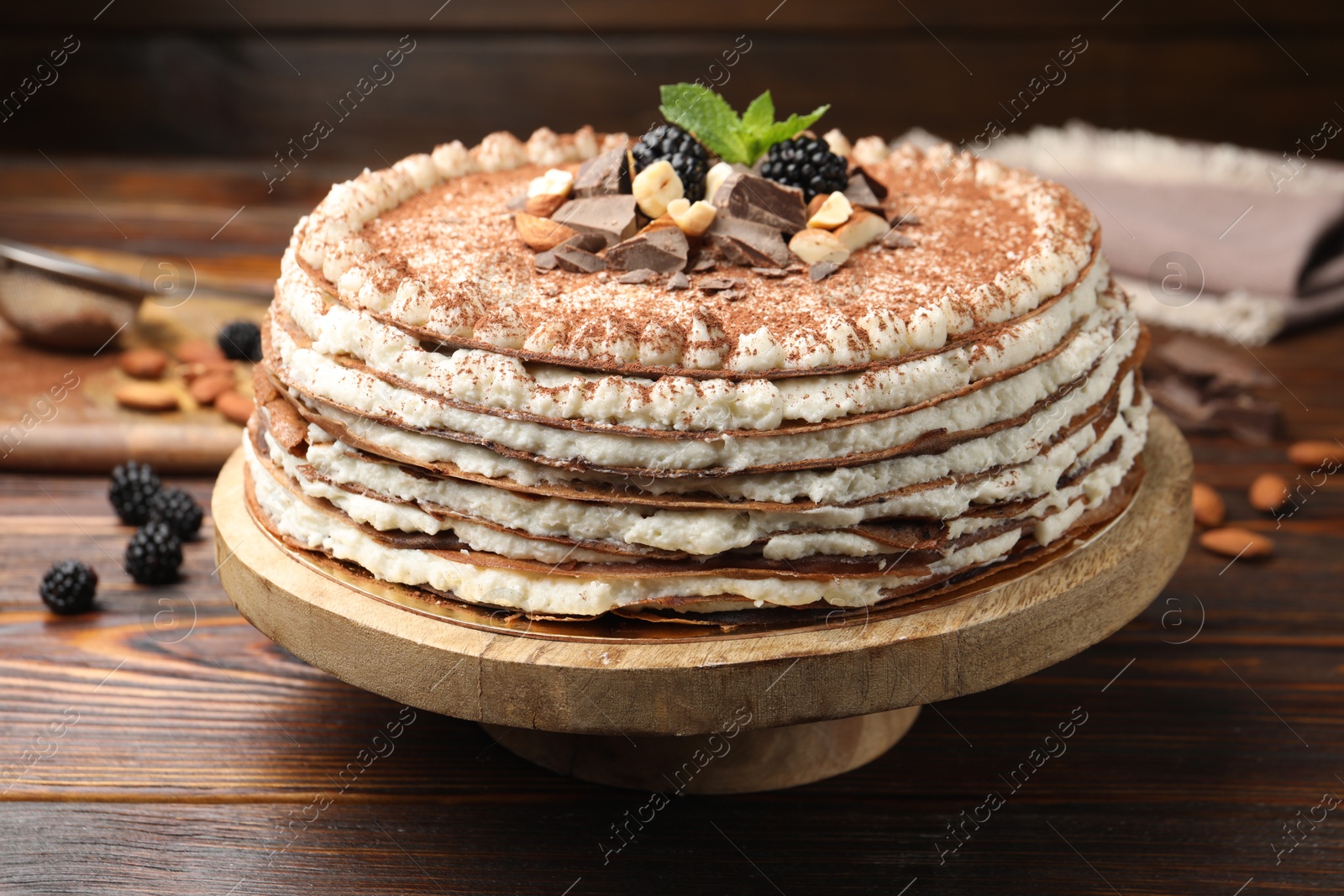 Photo of Delicious crepe cake with chocolate, nuts, blackberries and cocoa powder on wooden table, closeup