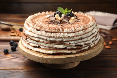 Photo of Delicious crepe cake with chocolate, nuts, blackberries and cocoa powder on wooden table, closeup