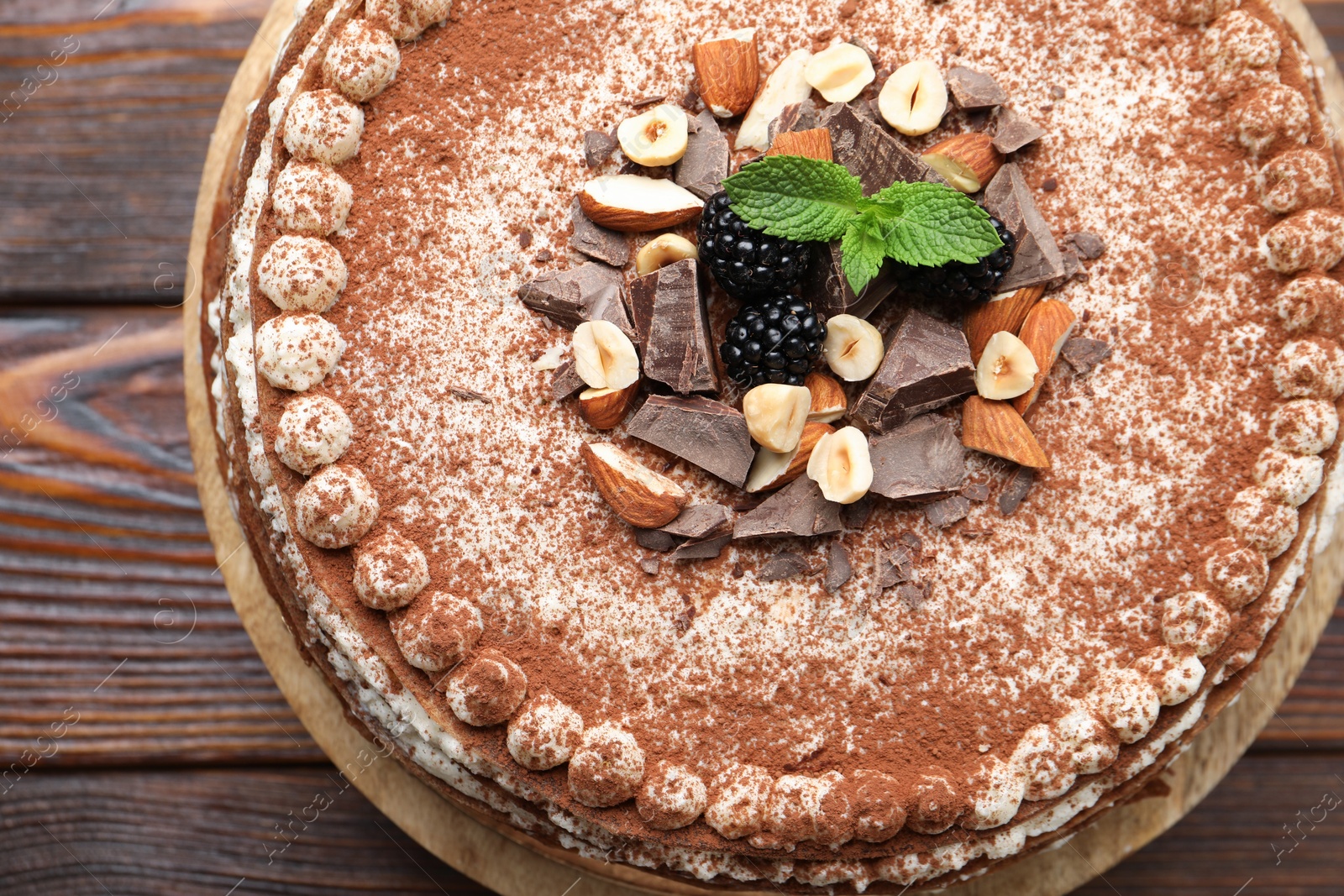 Photo of Delicious crepe cake with chocolate, nuts, blackberries and cocoa powder on wooden table, top view