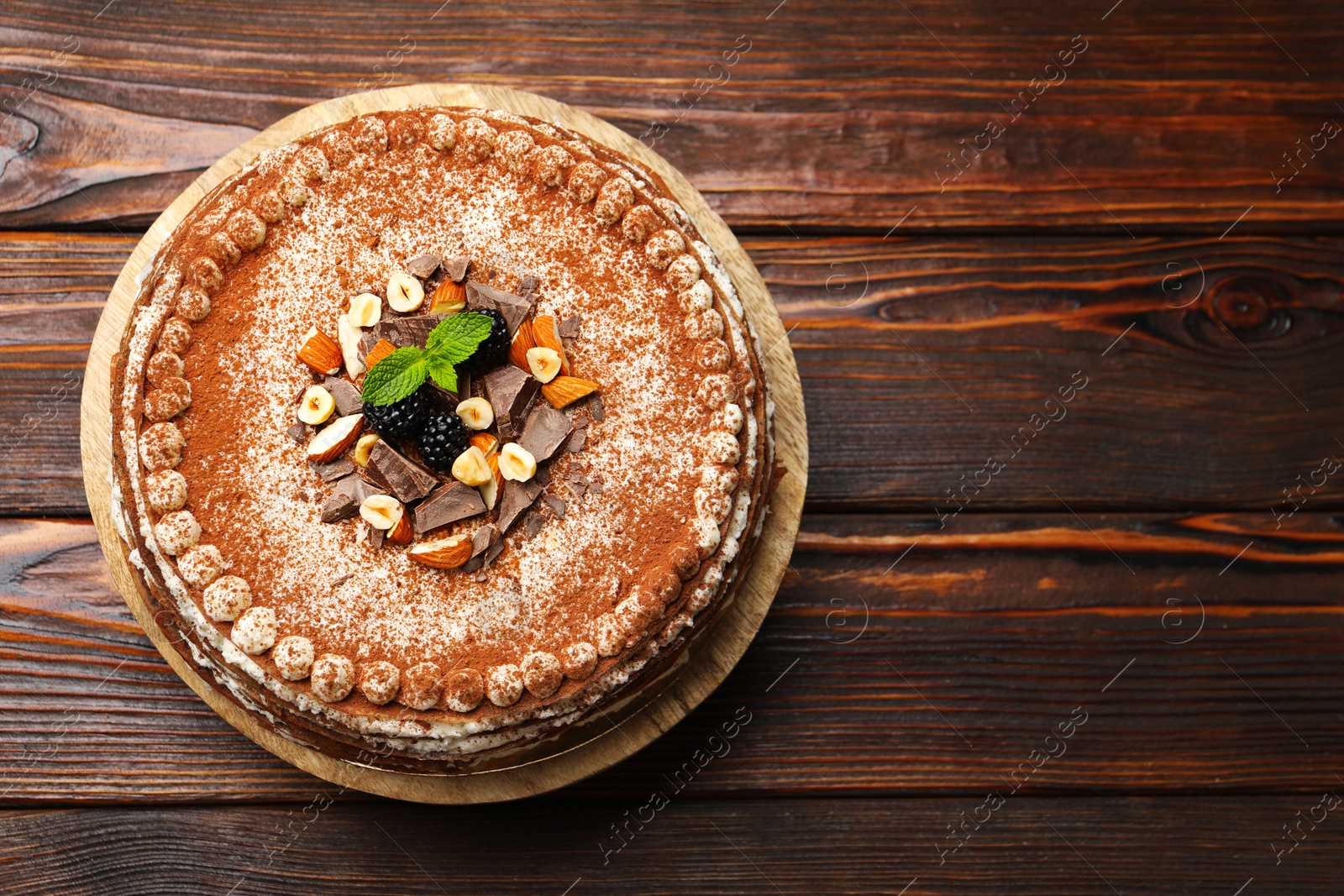 Photo of Delicious crepe cake with chocolate, nuts, blackberries and cocoa powder on wooden table, top view. Space for text