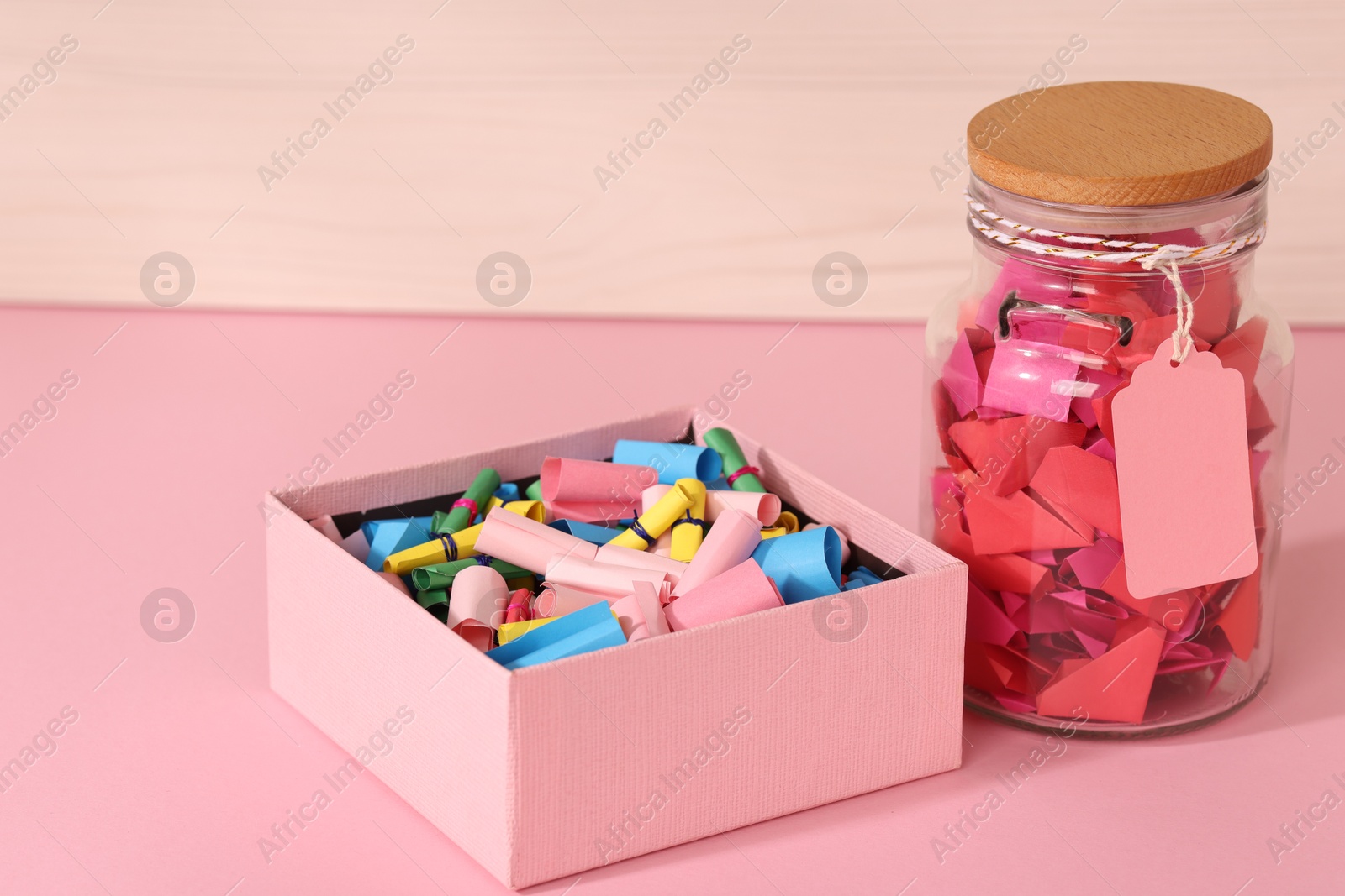 Color paper pieces in box and glass jar on pink table Photo of Color paper pieces in box and glass jar on pink table