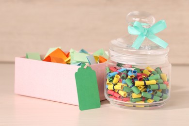 Photo of Color paper pieces in box and glass jar on light wooden table, closeup