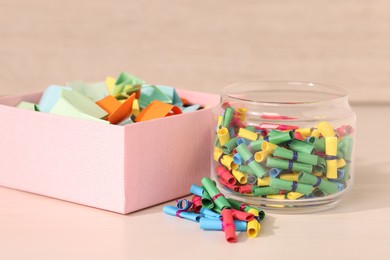 Photo of Color paper pieces in box and glass jar on light wooden table, closeup