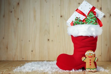 Photo of Saint Nicholas day tradition. Red boot with sweets, gift box and artificial snow on wooden table, space for text