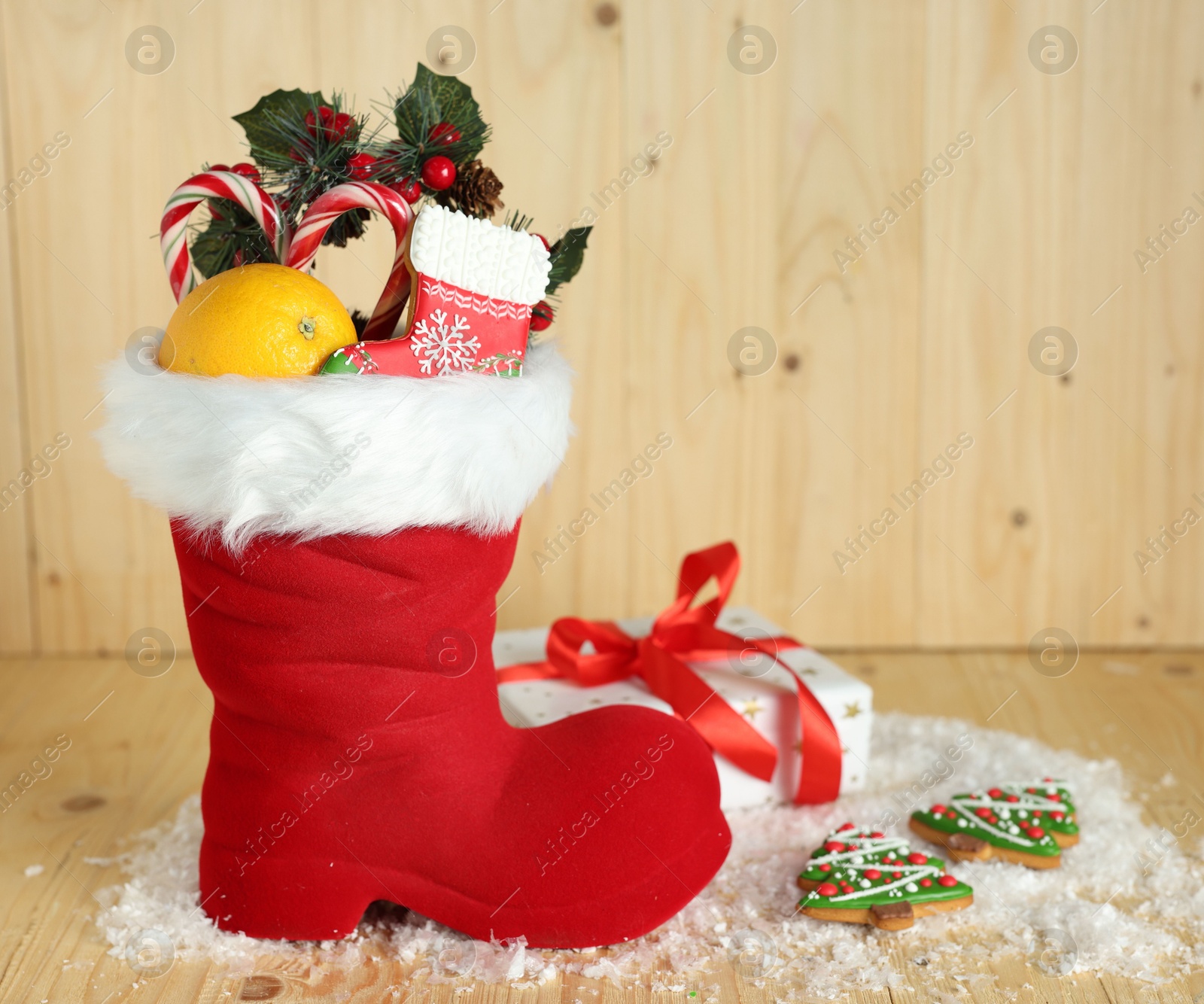 Photo of Saint Nicholas day tradition. Red boot with sweets, festive decor, gift box and artificial snow on wooden table