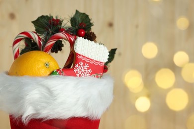 Photo of Saint Nicholas day tradition. Red boot with sweets and festive decor against wooden background with blurred lights, closeup. Space for text