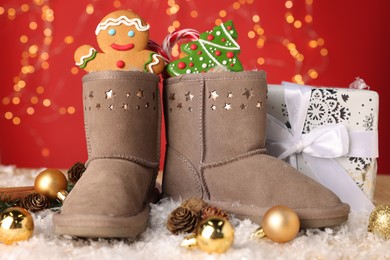 Photo of Saint Nicholas day. Boots filled with treats, festive decor and gift box on table against red background with blurred lights. Bokeh effect