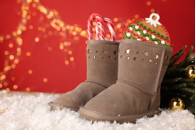 Photo of Saint Nicholas day. Boots filled with treats and festive decor on artificial snow against red background with blurred lights, space for text. Bokeh effect