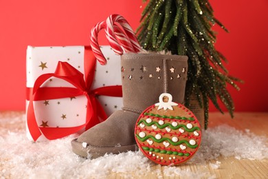 Photo of Saint Nicholas day. Boot with treats, gift box, fir tree and artificial snow on wooden table against red background