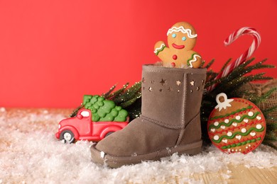 Photo of Saint Nicholas day. Boot with treats, gift box, fir tree and artificial snow on wooden table against red background