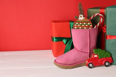 Photo of Saint Nicholas day. Boot with treats, fir branch and gift boxes on white wooden table against red background. Space for text