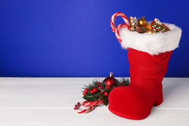 Photo of Saint Nicholas day. Red boot with treats and festive decor on white wooden table against blue background. Space for text