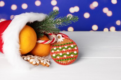 Photo of Saint Nicholas day. Boot with treats and fir branch on white wooden table against blue background with blurred lights, space for text. Bokeh effect
