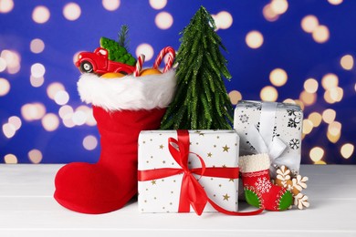 Photo of Saint Nicholas day. Red boot with treats, gift boxes and fir tree on white wooden table against blue background with blurred lights, bokeh effect