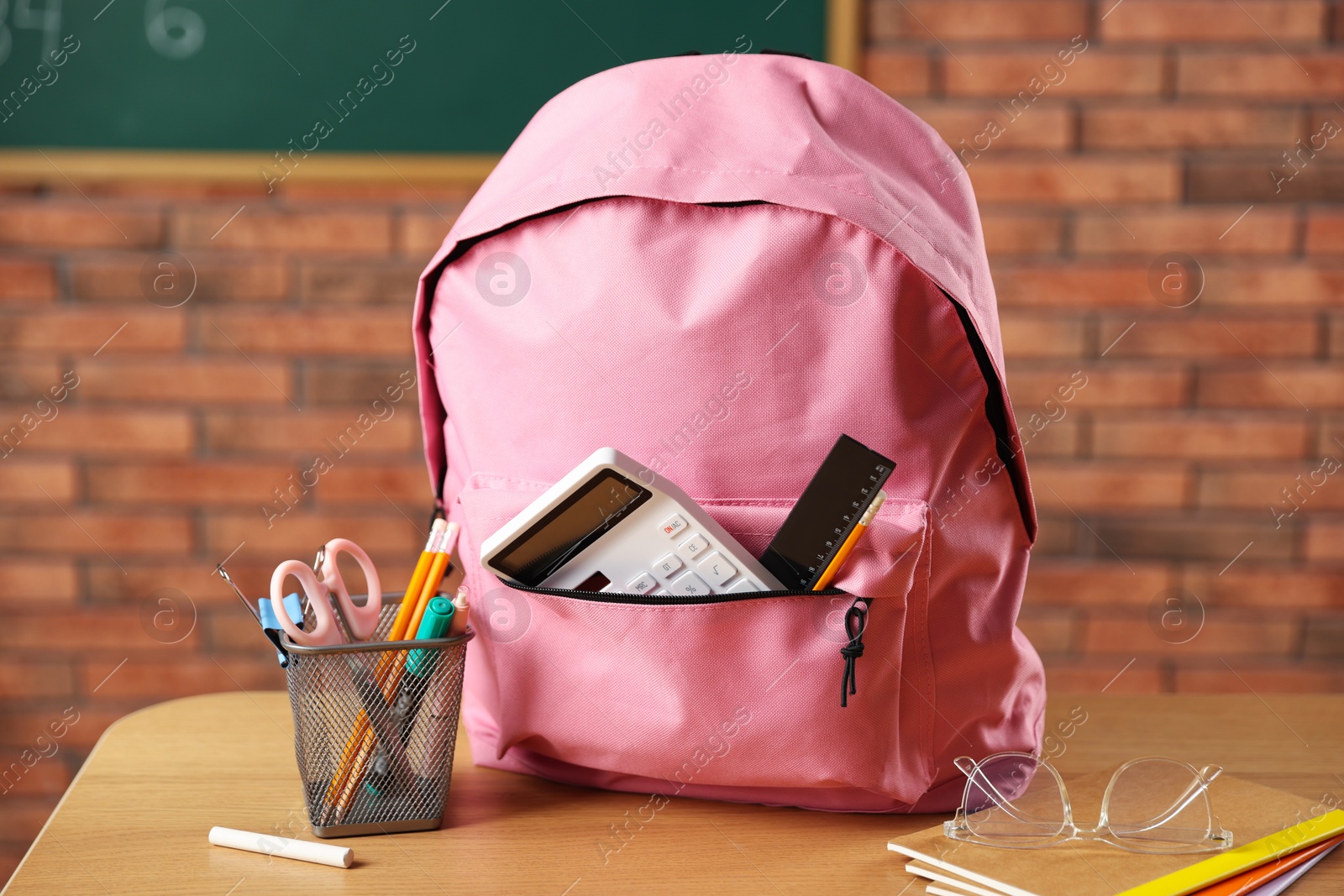 Pink backpack with different stationery on desk in classroom Photo of Pink backpack with different stationery on desk in classroom