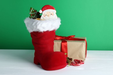 Photo of Happy Saint Nicholas day. Red boot with sweets and gift box on white wooden table against green background