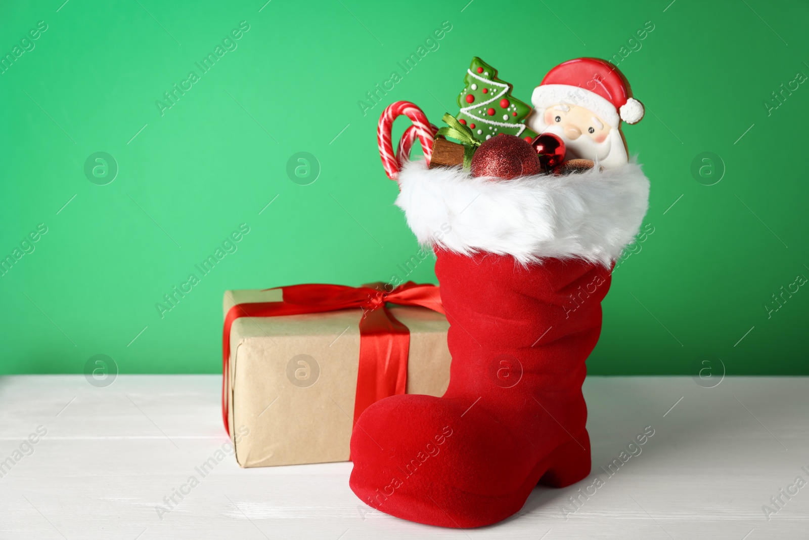 Happy Saint Nicholas day. Red boot with sweets, festive decor and gift box on white wooden table against green background Photo of Happy Saint Nicholas day. Red boot with sweets, festive decor and gift box on white wooden table against green background