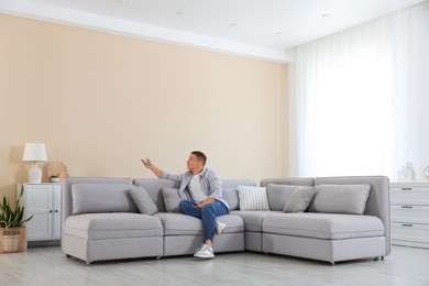 Photo of Man using remote to control air conditioner on sofa at home