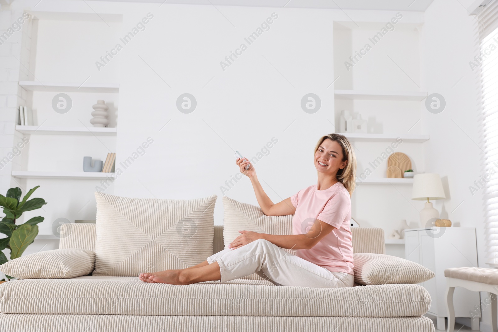 Photo of Woman using remote to control air conditioner on sofa at home