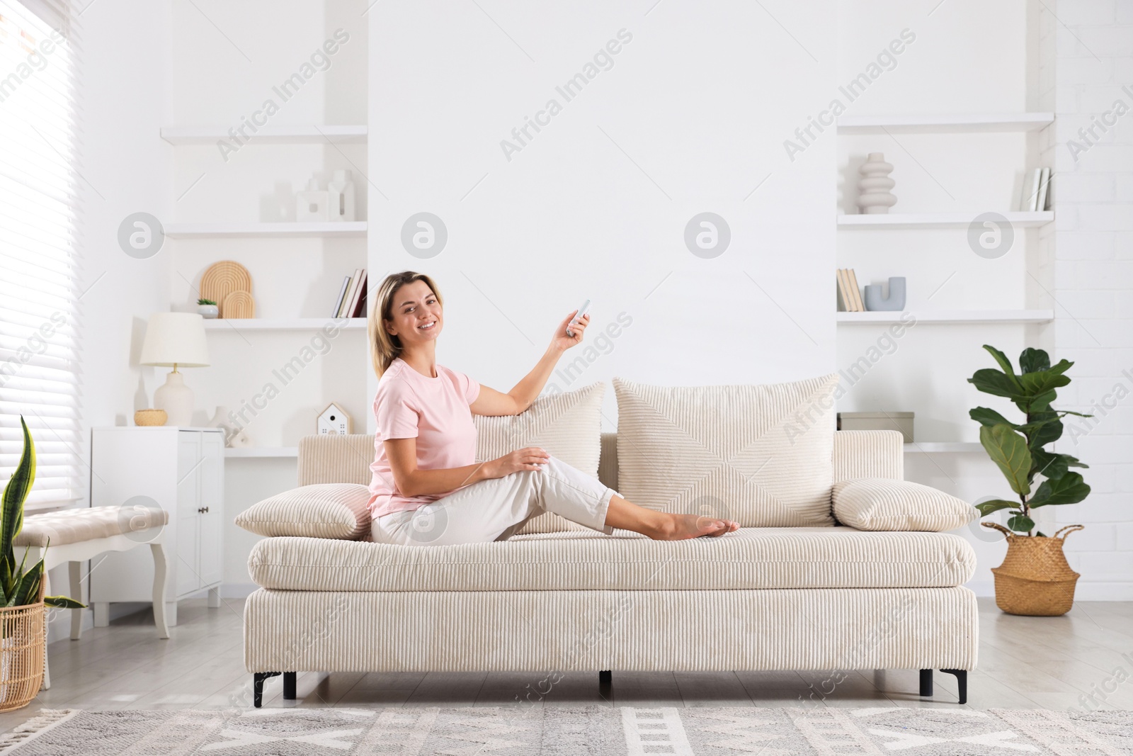 Photo of Woman using remote to control air conditioner on sofa at home