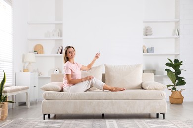 Photo of Woman using remote to control air conditioner on sofa at home