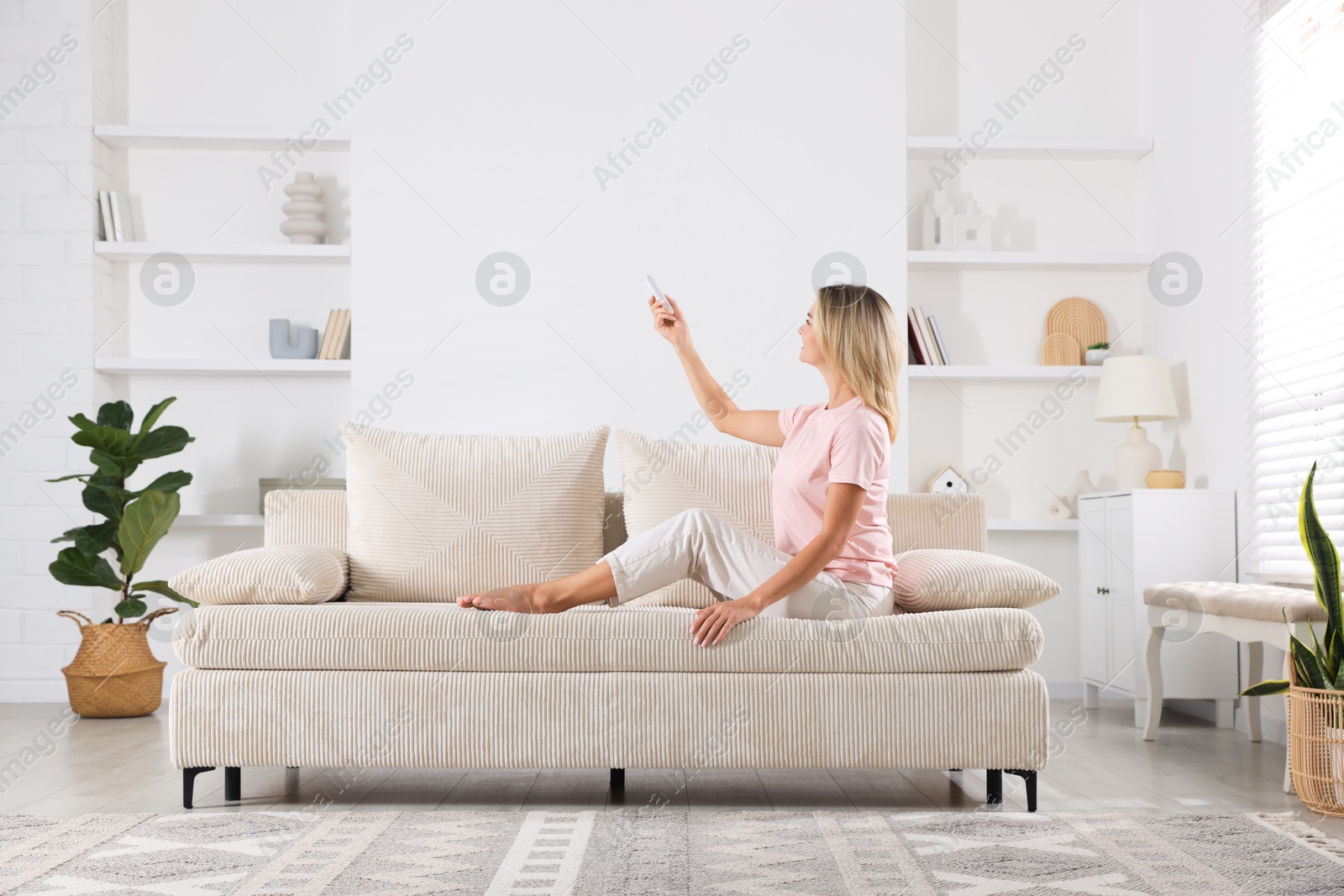 Photo of Woman using remote to control air conditioner on sofa at home