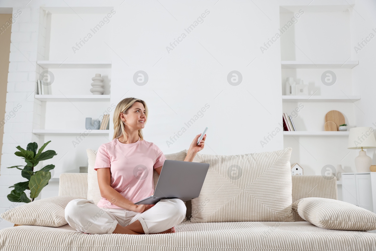 Photo of Woman with laptop using remote to control air conditioner on sofa at home