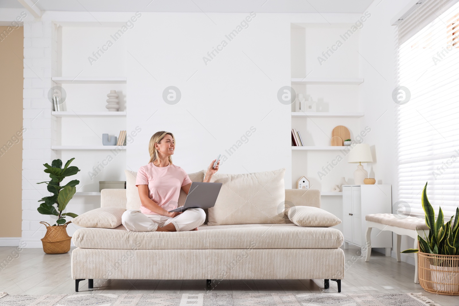 Photo of Woman with laptop using remote to control air conditioner on sofa at home