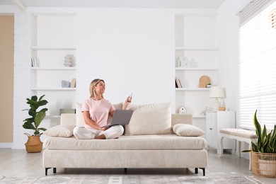Photo of Woman with laptop using remote to control air conditioner on sofa at home