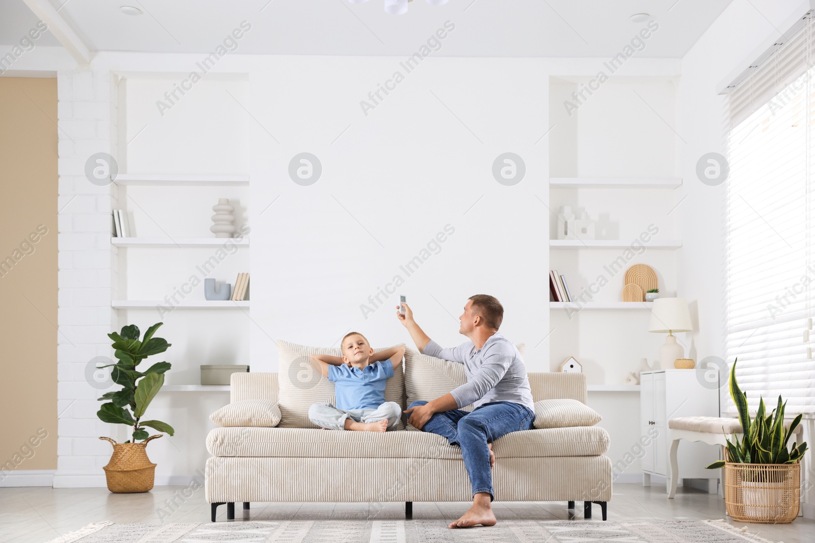 Photo of Cute little boy and his father using remote to control air conditioner on sofa at home