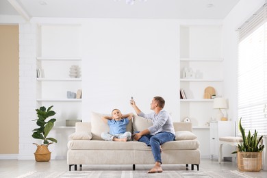 Photo of Cute little boy and his father using remote to control air conditioner on sofa at home